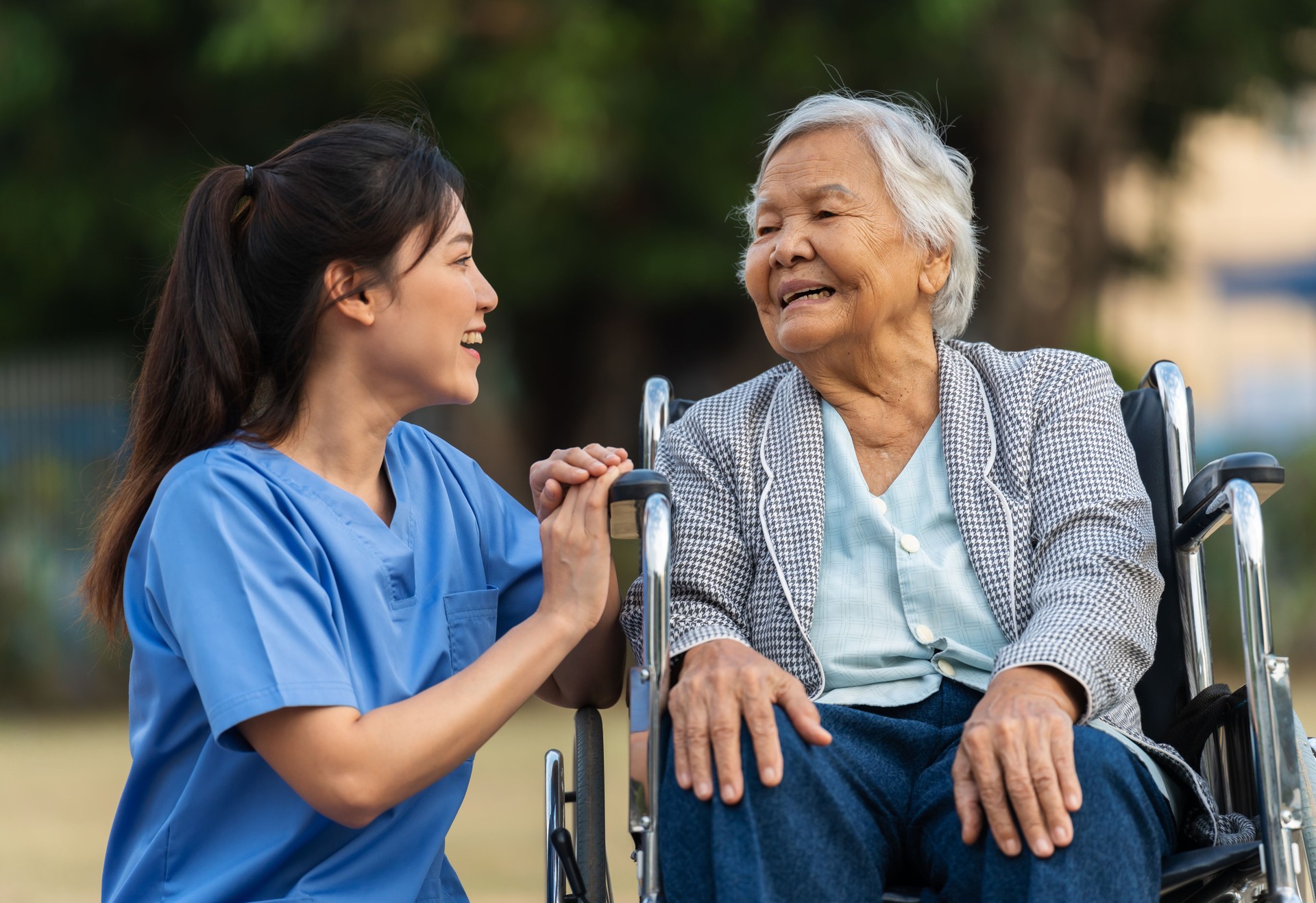 happy nurse take care and talking with senior woman patient in wheelchair at park, friendly caregiver